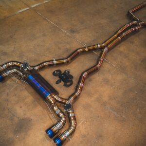 Shiny aftermarket car exhaust pipes laid out on a concrete workshop floor, featuring blue-tipped mufflers and mounting hardware.