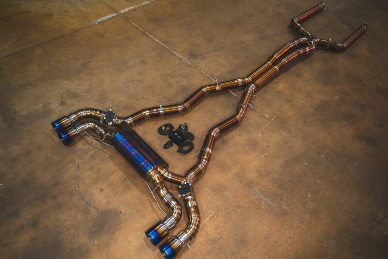A7401071jpg - OPSHOLDERS Shiny aftermarket car exhaust pipes laid out on a concrete workshop floor, featuring blue-tipped mufflers and mounting hardware.