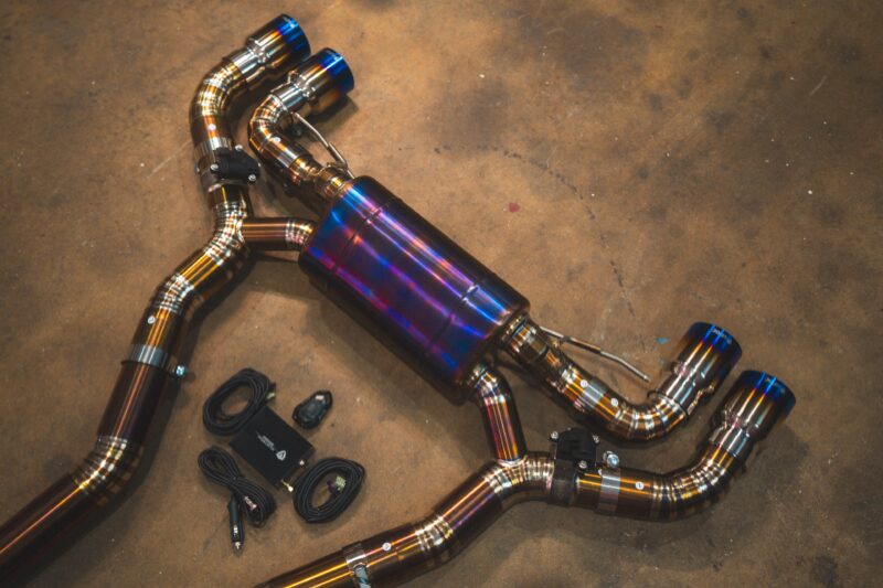 A7401072jpg - OPSHOLDERS Colorful titanium exhaust pipes arranged on a concrete workshop floor, with connectors and cables nearby, forming an intricate curved layout.