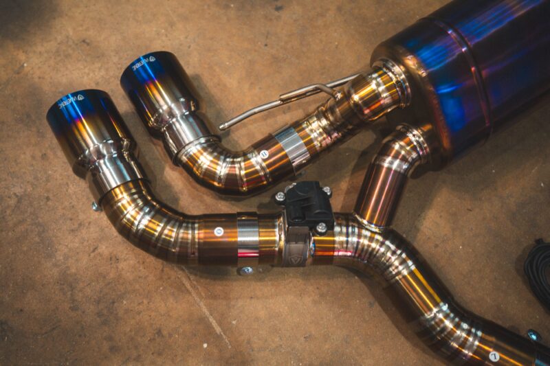 A7401075jpg - OPSHOLDERS Close-up of a polished stainless steel exhaust system with curved pipes on a concrete workshop floor.