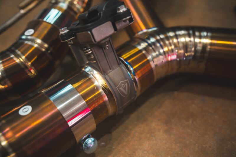 A7401077jpg - OPSHOLDERS Close-up of metallic pipes and a black valve clamp, with reflective copper tubing in an industrial setting.