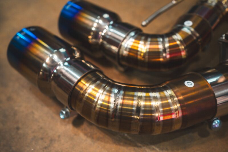 A7401079jpg - OPSHOLDERS Close-up of polished stainless steel exhaust pipes curved in a circular arrangement on a workshop floor.