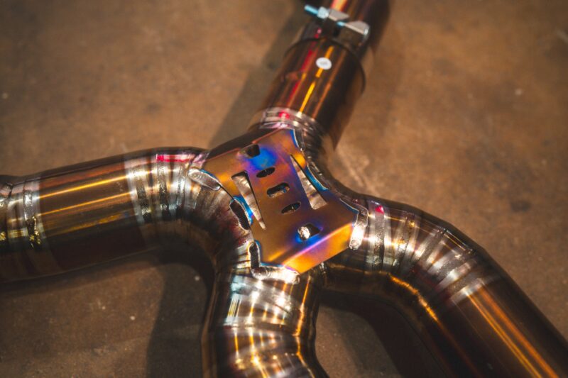 A7401080jpg - OPSHOLDERS Close-up of metallic welded joints on curved chrome tubes with rainbow heat-color patina forming an intersection.