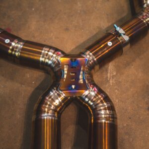 Welded stainless steel exhaust pipes arranged in a cross on a concrete floor with clamps and a central hub.