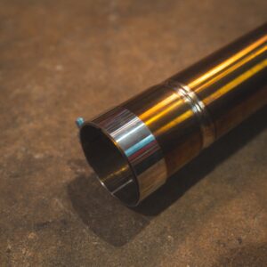 Close-up of a polished metal exhaust pipe resting on a rough concrete workshop floor, with reflections visible on the pipe’s end cap and welds nearby.
