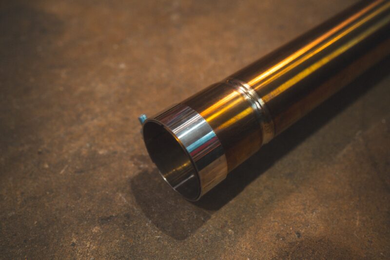 A7401082jpg - OPSHOLDERS Close-up of a polished metal exhaust pipe resting on a rough concrete workshop floor, with reflections visible on the pipe’s end cap and welds nearby.