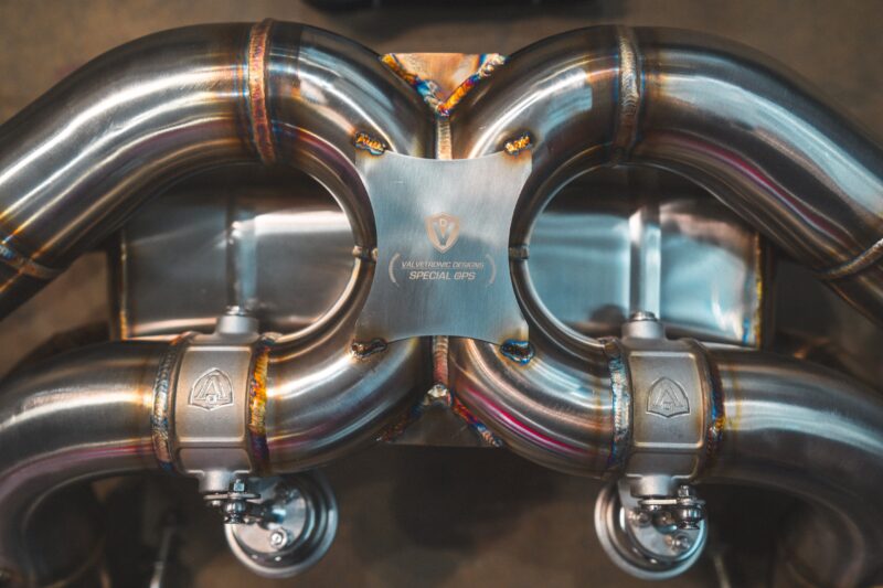 Close-up of polished stainless steel exhaust headers with welded joints and a central plaque bearing Valvetronic Designs Special OPS logo.