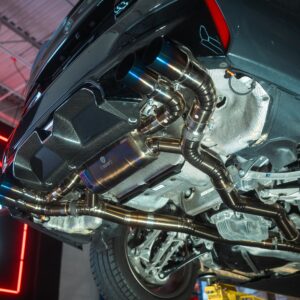 Underside of a dark sports car on a lift, showing polished exhaust pipes and heat shielding in a workshop light.