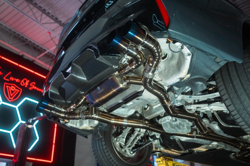 A7407920jpg - OPSHOLDERS Underside of a dark sports car on a lift, showing polished exhaust pipes and heat shielding in a workshop light.