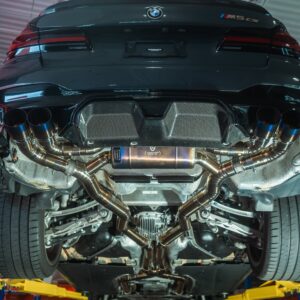 Underside of a dark BMW M5 with exposed dual exhausts in a workshop, car raised on a lift with red shelves in the background.