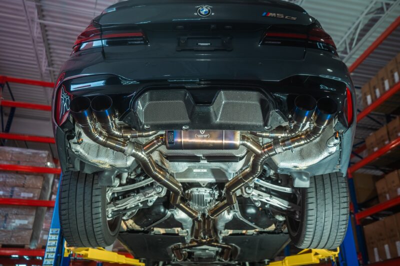 A7407921jpg - OPSHOLDERS Underside of a dark BMW M5 with exposed dual exhausts in a workshop, car raised on a lift with red shelves in the background.