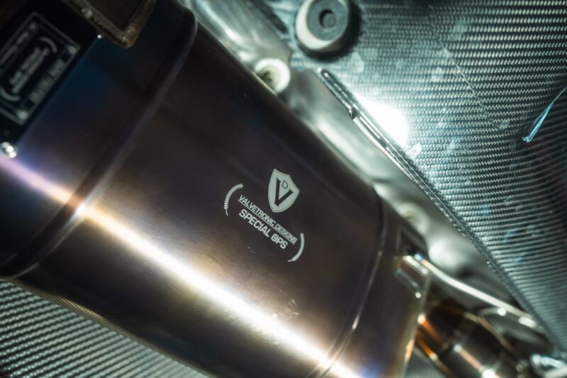 A7407922jpg - OPSHOLDERS Close-up of a stainless steel exhaust pipe with Valvetronic Designs Special Ops branding, against a carbon-fiber background.