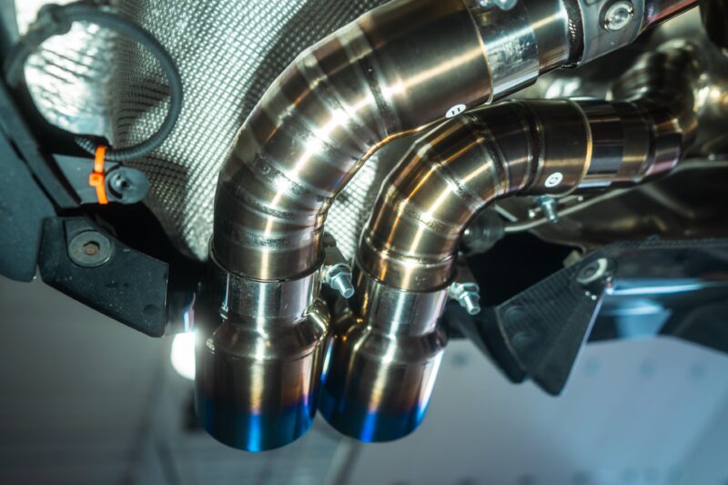 A7407924jpg - OPSHOLDERS Close-up of shiny curved stainless steel exhaust pipes and clamps in a vehicle engine bay.