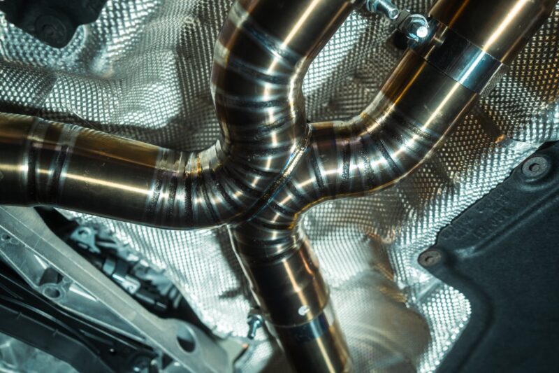 A7407928jpg - OPSHOLDERS Close-up of interconnected shiny stainless steel exhaust pipes with a textured heat shield in a car's engine bay