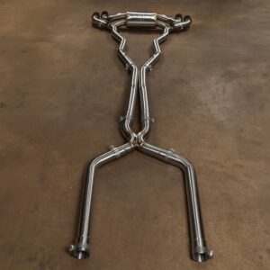 Polished stainless steel car exhaust pipes laid out on a brown workshop floor, arranged in a humanoid shape with arms raised and a central muffler at the top.