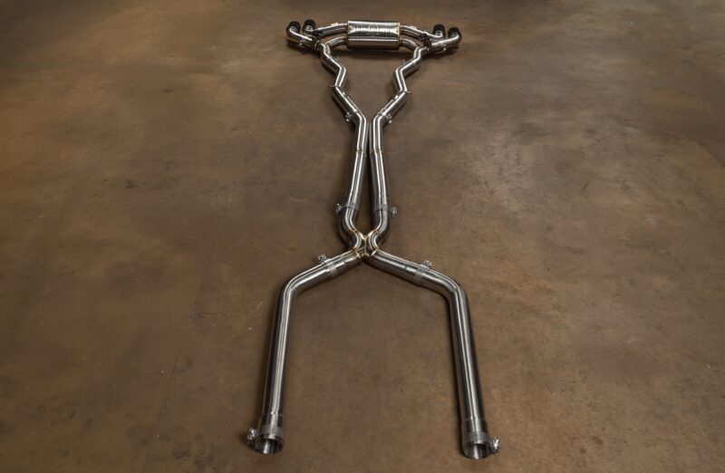 BMWF9XM5M8VSES_BR_RC_3jpg - OPSHOLDERS Polished stainless steel car exhaust pipes laid out on a brown workshop floor, arranged in a humanoid shape with arms raised and a central muffler at the top.