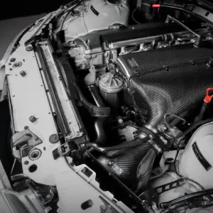 Close-up of a car engine bay in grayscale, highlighting a carbon-fiber intake cover, hoses, and surrounding metal components.