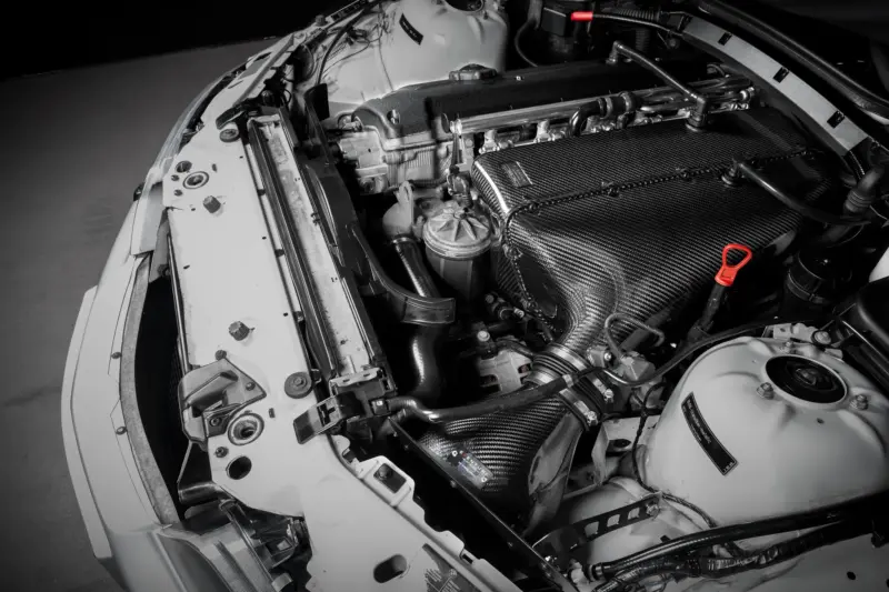 Close-up of a car engine bay in grayscale, highlighting a carbon-fiber intake cover, hoses, and surrounding metal components.