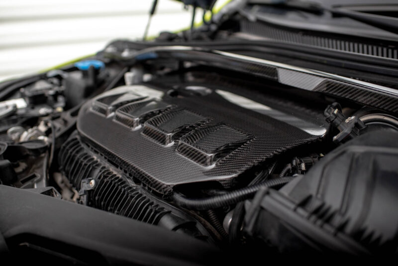 Close-up of a car engine bay showcasing carbon fiber panels and nearby wiring/hoses under the hood.