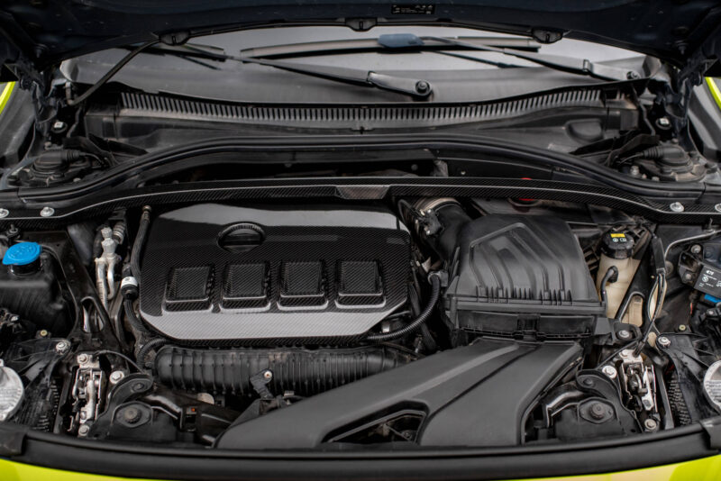 Front view of a car engine bay with carbon fiber components and a blue radiator cap visible on the left side.