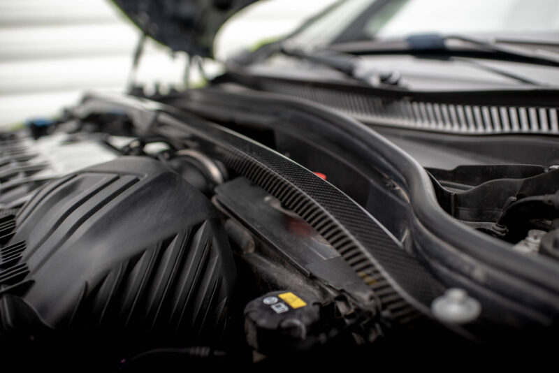 Under-hood view of a car engine bay with hoses and engine components visible in the foreground and background out of focus.