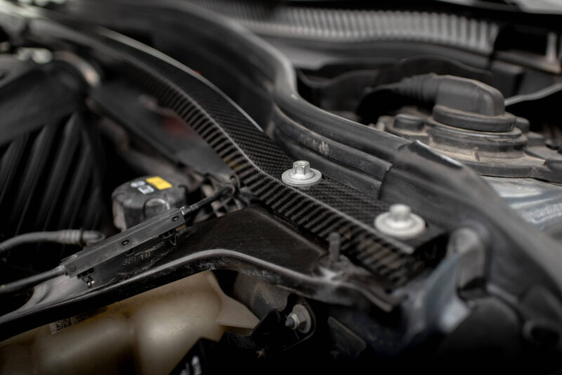 Close-up of an automotive engine bay with hoses, a coolant reservoir, and a metal bolt in focus under dim lighting.