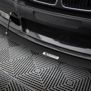Close-up of a black BMW front bumper with an aftermarket lip and a vertical support rod, on a patterned metal floor surface.