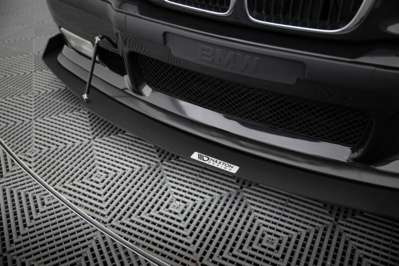 Close-up of a black BMW front bumper with an aftermarket lip and a vertical support rod, on a patterned metal floor surface.