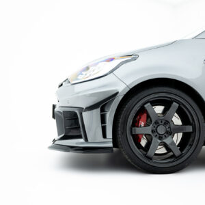 Close-up of the front left side of a gray sports car showing the headlight, fender badge, and black alloy wheel with a red brake caliper.