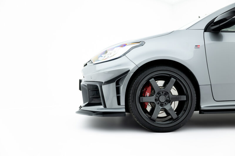 Close-up of the front left side of a gray sports car showing the headlight, fender badge, and black alloy wheel with a red brake caliper.