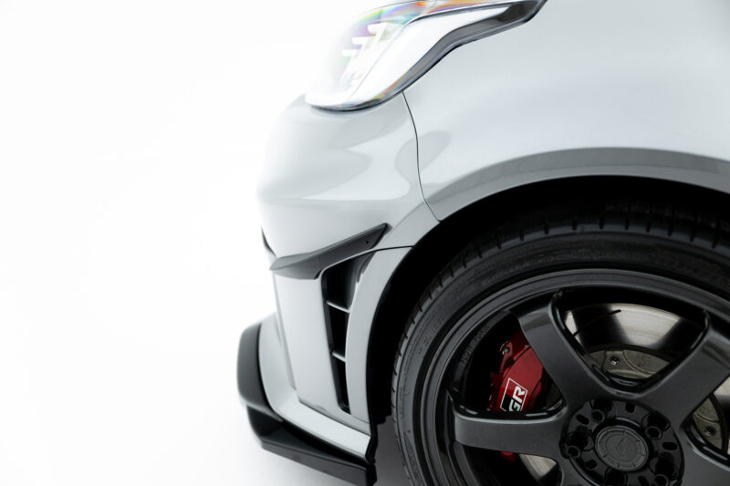 Close-up of a white car's front left corner showing the headlight and black wheel with red brake caliper behind it