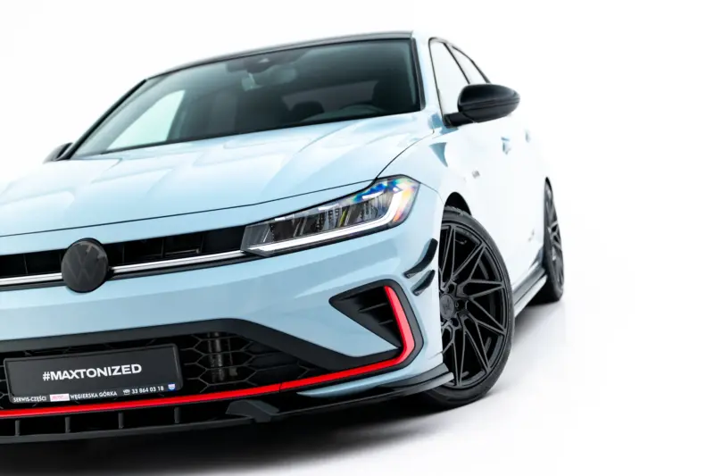 Front-left angled view of a light-blue Volkswagen car showing the headlight, emblem, and large black alloy wheel with a red accent. Plate reads #MAXTONIZED.