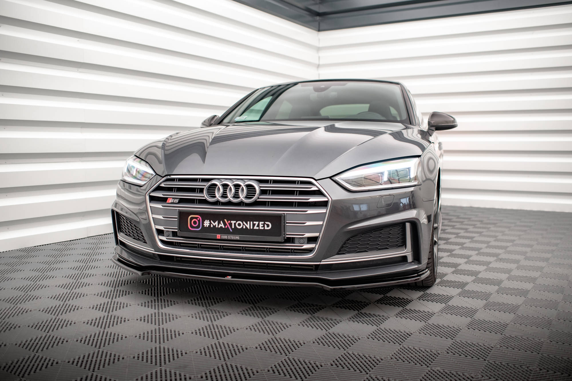 pol_po_Front-Splitter-V-1-Audi-S5-A5-S-Line-F5-Coupe-Sportback-5599_4jpg - OPSHOLDERS Front three-quarter view of a grey Audi sedan in a showroom, with a black plate reading #MAXTONIZED at the front.