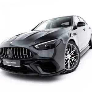 Close-up of a dark gray Mercedes-AMG car, showing the front grille, headlight, and large alloy wheels on a white background, angled from the left-front corner.