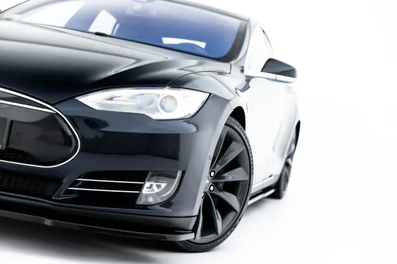 Close-up of a glossy black car front, featuring the headlight, bumper, and wheel in view.