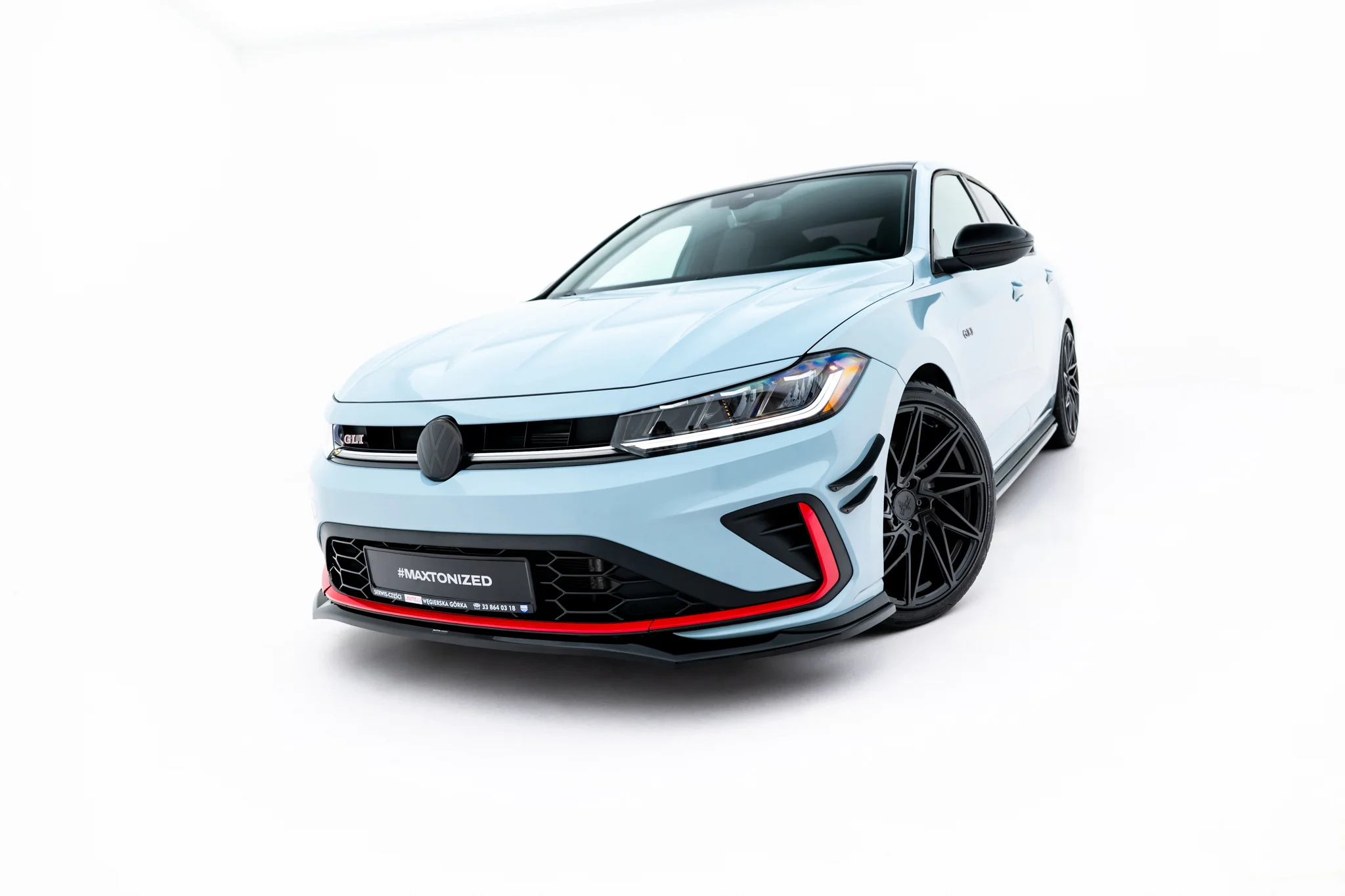 pol_po_Front-Splitter-V-2-Volkswagen-Jetta-GLI-Mk7-Facelift-2-23930_6webp - OPSHOLDERS Front-left close-up of a light-blue sporty car in a white studio, highlighting bold front grille and large black wheels with red accents on the bumper badge reading '#MAXTONIZED'.