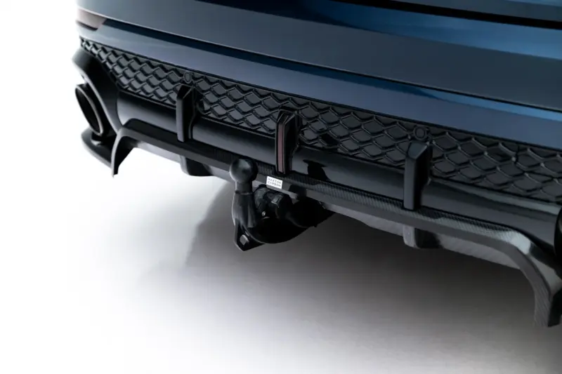 Close-up of a blue car's front bumper showing a honeycomb grille and carbon fiber trim with a towing hitch component visible below.
