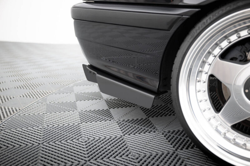 pol_po_REAR-DIFFUSER-BMW-M3-3-M-Pack-Coupe-Cabrio-E36-1829_11jpg - OPSHOLDERS Close-up of a black car bumper and chrome wheel on a textured showroom floor.