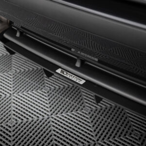 Close-up of the rear of a dark car showing dual exhaust tips and a Maxton Design badge on a textured floor surface.