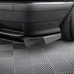 Close-up of a glossy black car’s front bumper and adjacent wheel with a large chrome rim, parked on a textured checkered floor.