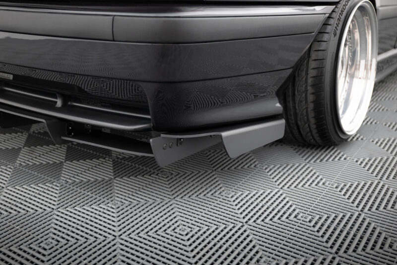 pol_po_REAR-DIFFUSER-BMW-M3-3-M-Pack-Coupe-Cabrio-E36-1829_9jpg - OPSHOLDERS Close-up of a glossy black car’s front bumper and adjacent wheel with a large chrome rim, parked on a textured checkered floor.