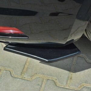 Close-up of a carbon fiber rear diffuser under a dark car, near a large tire on a paved surface.