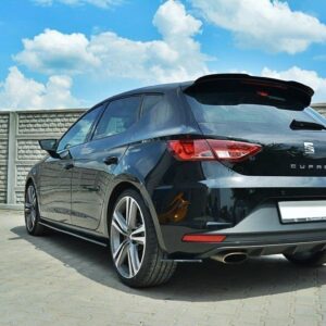 Rear three-quarter view of a black SEAT Cupra hatchback parked on a paved lot against a blue sky and concrete fence, showing the taillights and dual exhausts.