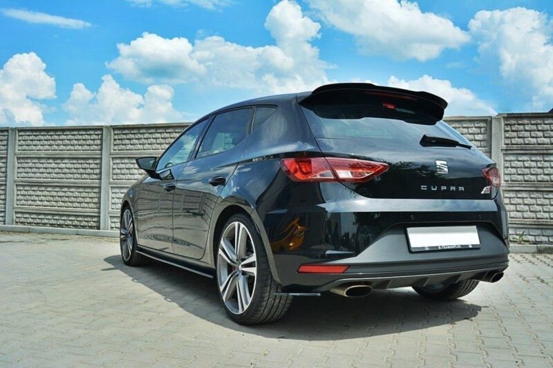 Rear three-quarter view of a black SEAT Cupra hatchback parked on a paved lot against a blue sky and concrete fence, showing the taillights and dual exhausts.