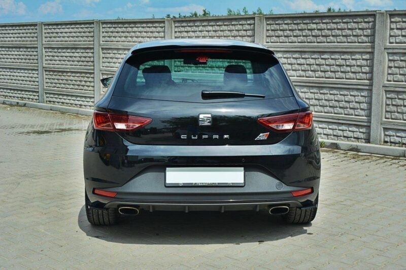Rear view of a black Seat Cupra hatchback parked on a paved surface with a tall concrete panel fence in the background.