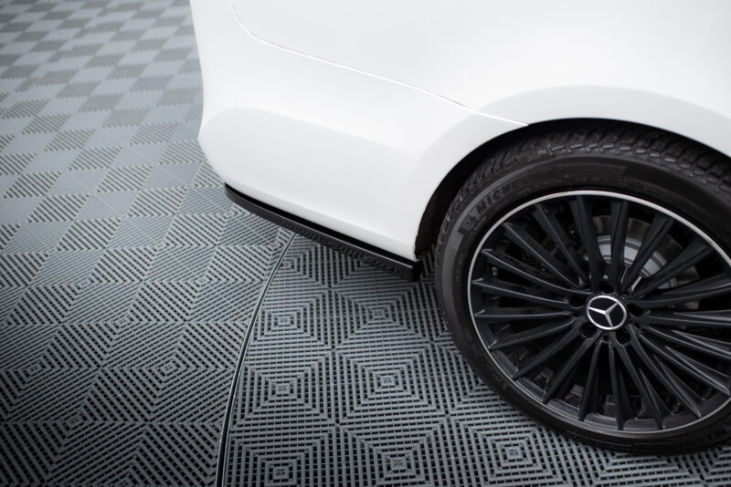 Close-up of a white car’s front fender and black alloy wheel on a checkered gray floor.