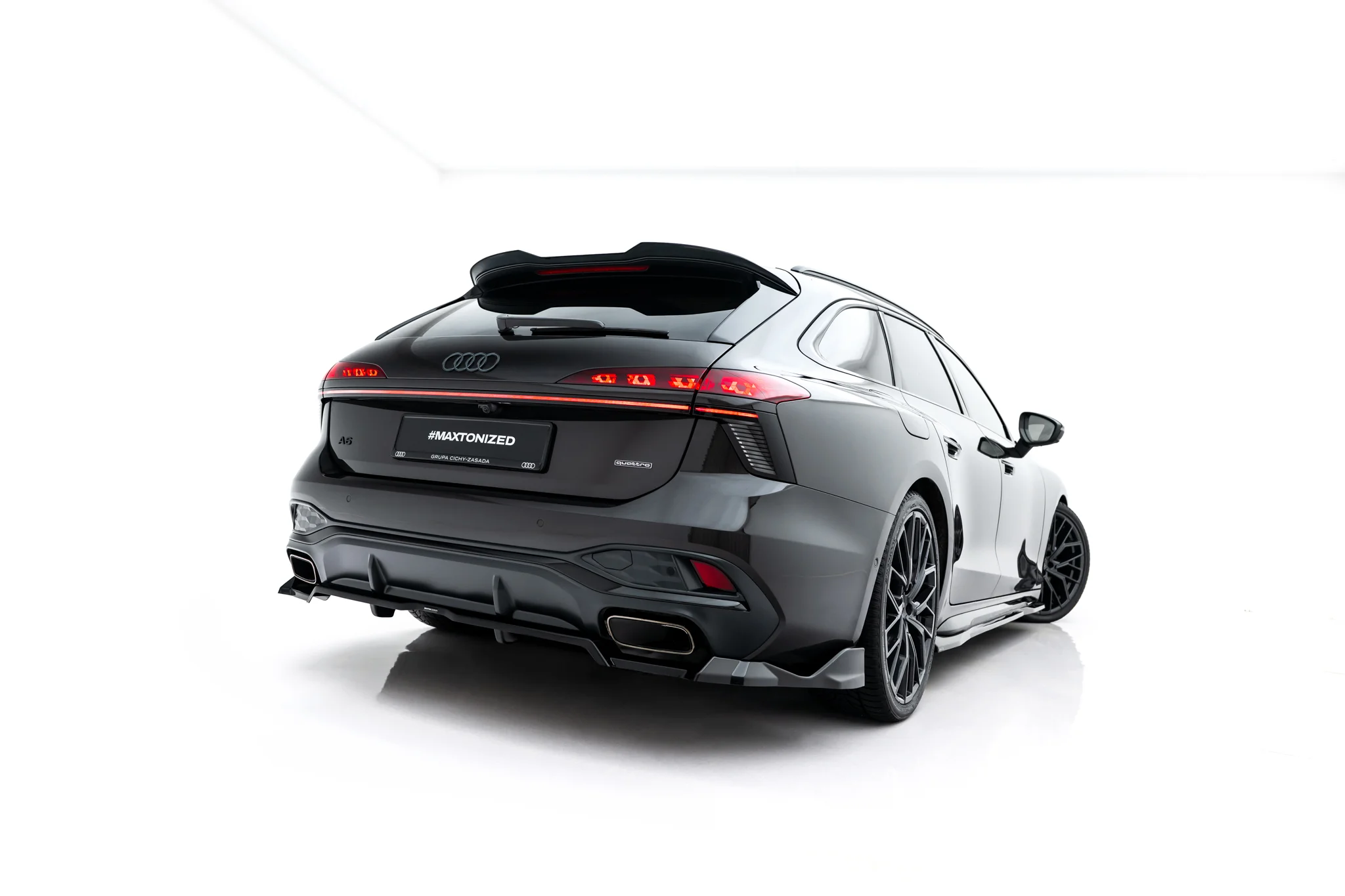 pol_po_Rear-Splitter-with-vertical-bars-V-4-Audi-A6-S-Line-C9-24007_4webp - OPSHOLDERS Rear three-quarter view of a dark Audi with illuminated tail lights, photographed against a white studio background.