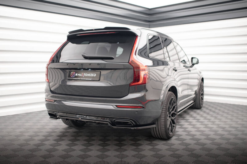 Rear three-quarter view of a dark gray Volvo SUV in a bright studio with striped walls; tail lights and license plate reading '#MAXTONIZED' visible.