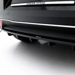 Close-up of a black car rear bumper with chrome trim and a Maxton Design badge visible along the lower edge, with soft left-side lighting and part of the exhaust circle in the corner.]