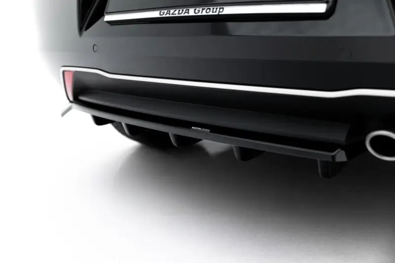Close-up of a black car rear bumper with chrome trim and a Maxton Design badge visible along the lower edge, with soft left-side lighting and part of the exhaust circle in the corner.]